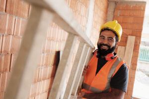 Confident people working in construction site. Portrait of happy hispanic man at work in new housing project. Professional latino worker using ladder and smiling at camera